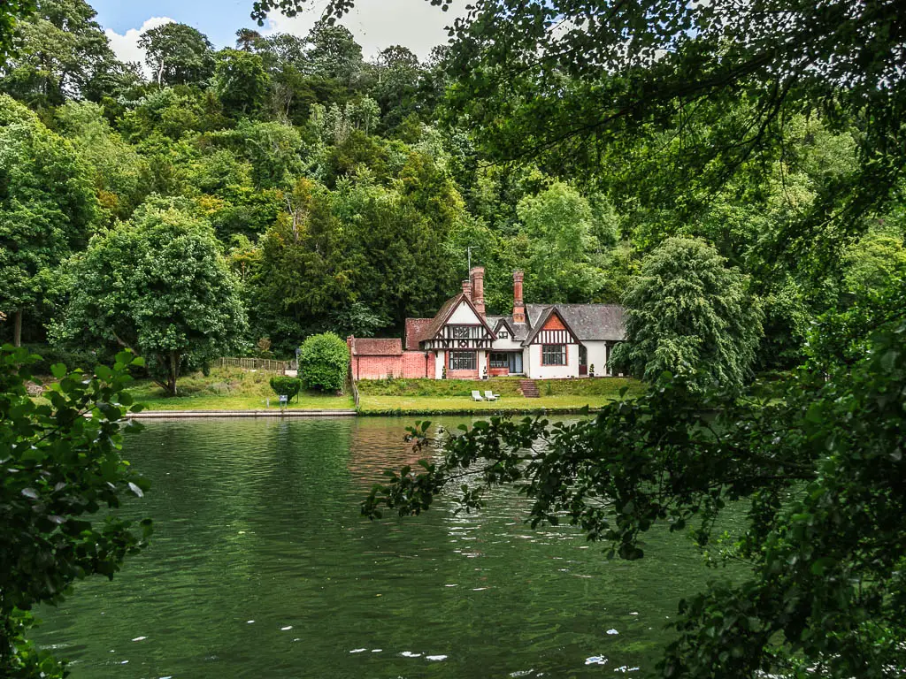 Looking through a gap in the trees, across the river to a Tudor house on the other side, near the end of the walk from Marlow to Maidenhead/ There is a neatly cut lawn in front of the hose and a mass of big green leafy trees behind it.