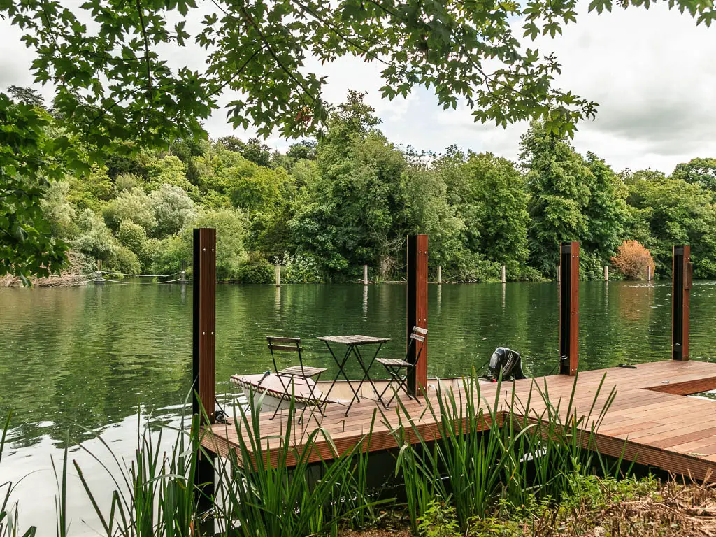 A wooden walking way on the edge of the river with a small table and two chairs on it, facing the river. There is a mass of green leafy trees on the other side of the river.