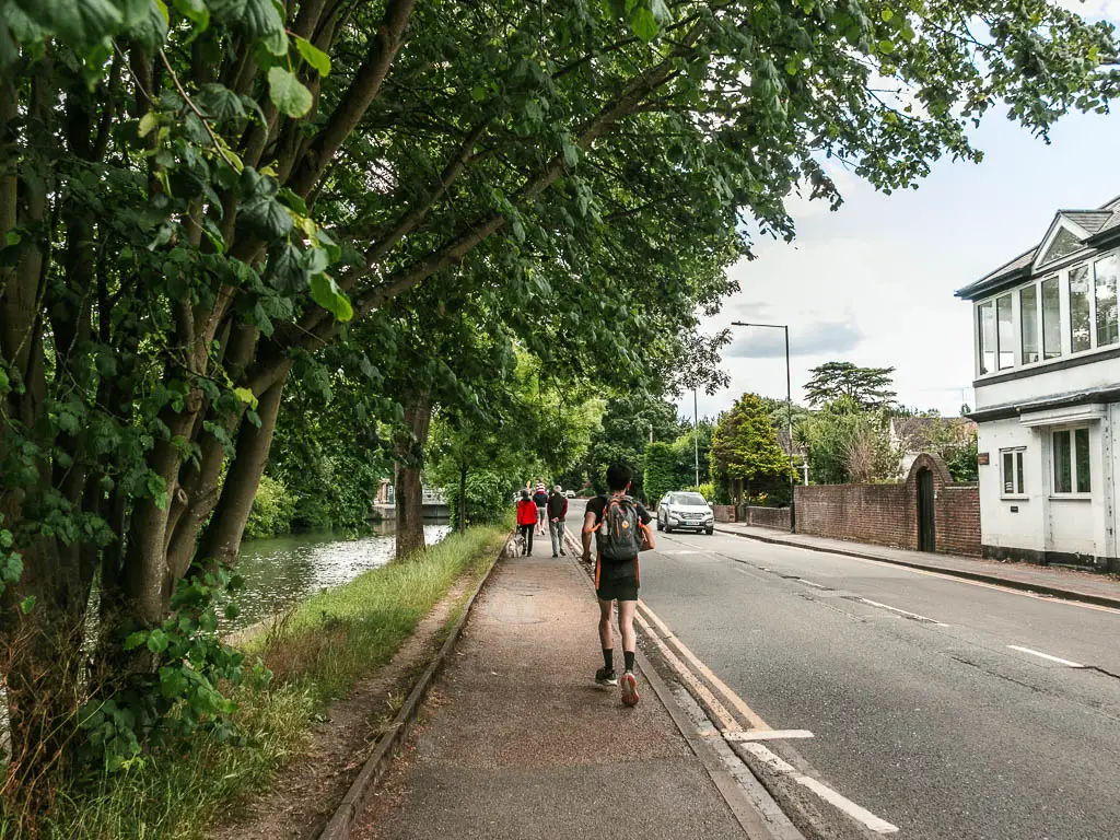 A pavement leading ahead, with the road too the right and strip of grass then the river to the left. There are people walking along the pavement. 