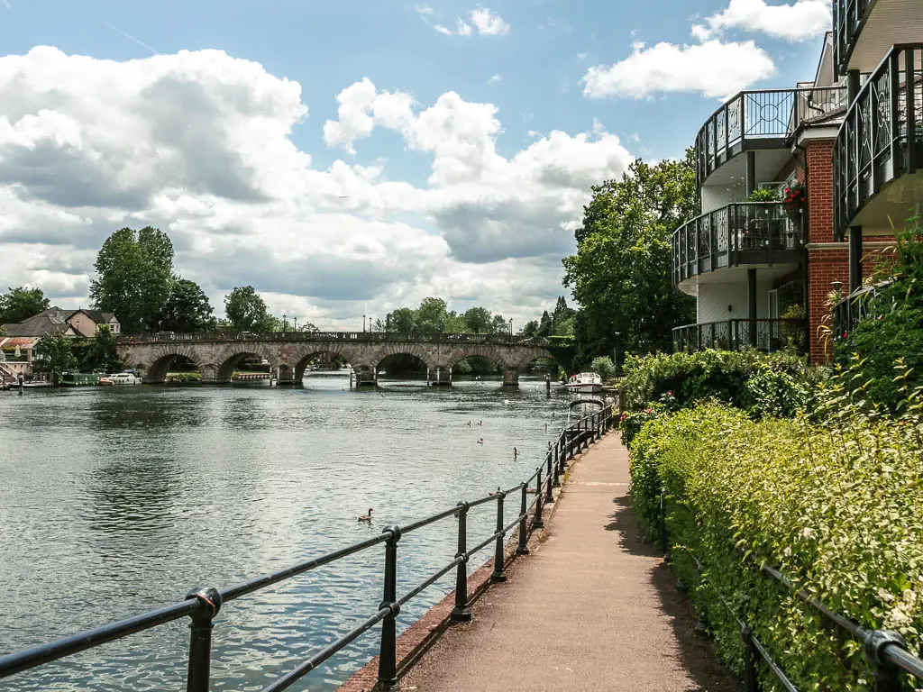 A walkway leading ahead, with metal railings and river on the left, and bushes and apartment buildings on the right. The archway maidenhead bridge is crossing the river ahead.