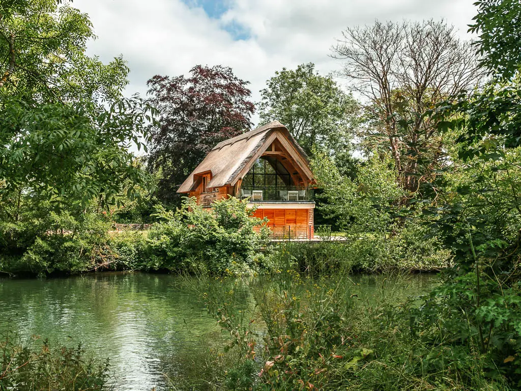 Looking over the bushes to the green coloured river and thatched roofed cottage on the other side, along the walk from Windsor to Staines. The cottage is surrounded by trees and bushes.