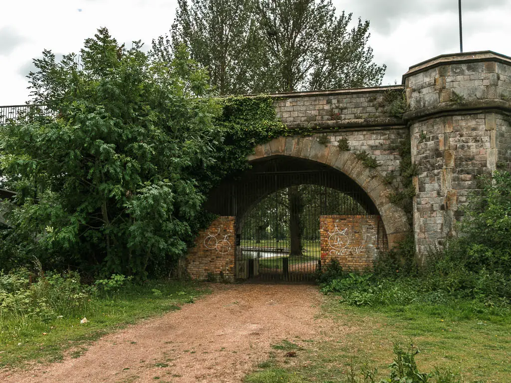 A wodę dirt trail leading to an arch under a stone bridler, and black metal gate blocking the trail.