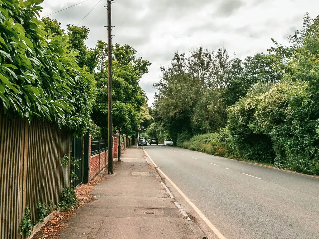 Looking along a pavement, with the road to the right and wooden fence to the left. Both sides of the road are lined with green leafy bushes and trees.