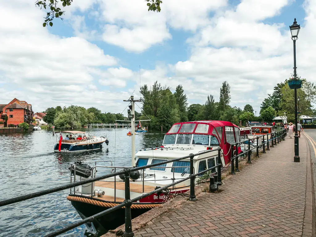 A walkway on the right, and river on the left with a black railing and boats moored to the side.