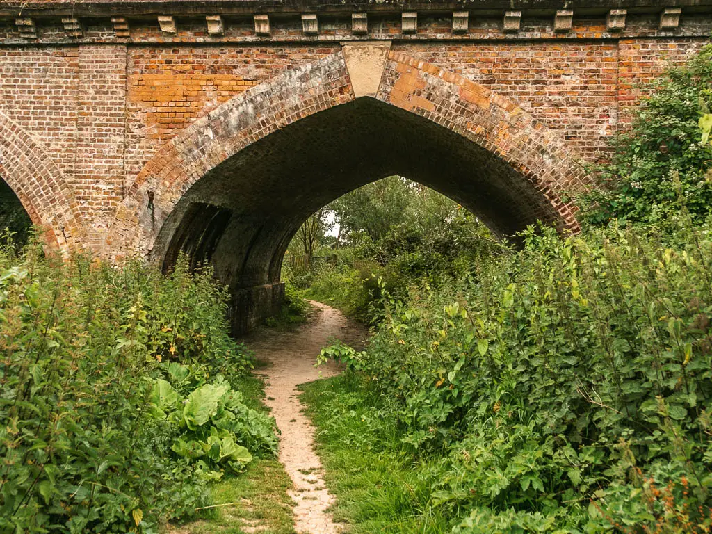 The trail lined with green bushes, leading under an archway of a big brick bridge.