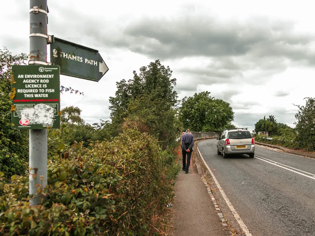 A small pavement leading ahead, with the road on the right and bushes on the left. There is a trail sign on the left side pointing along the road. There is a person walking on the pavement and a car on the road.