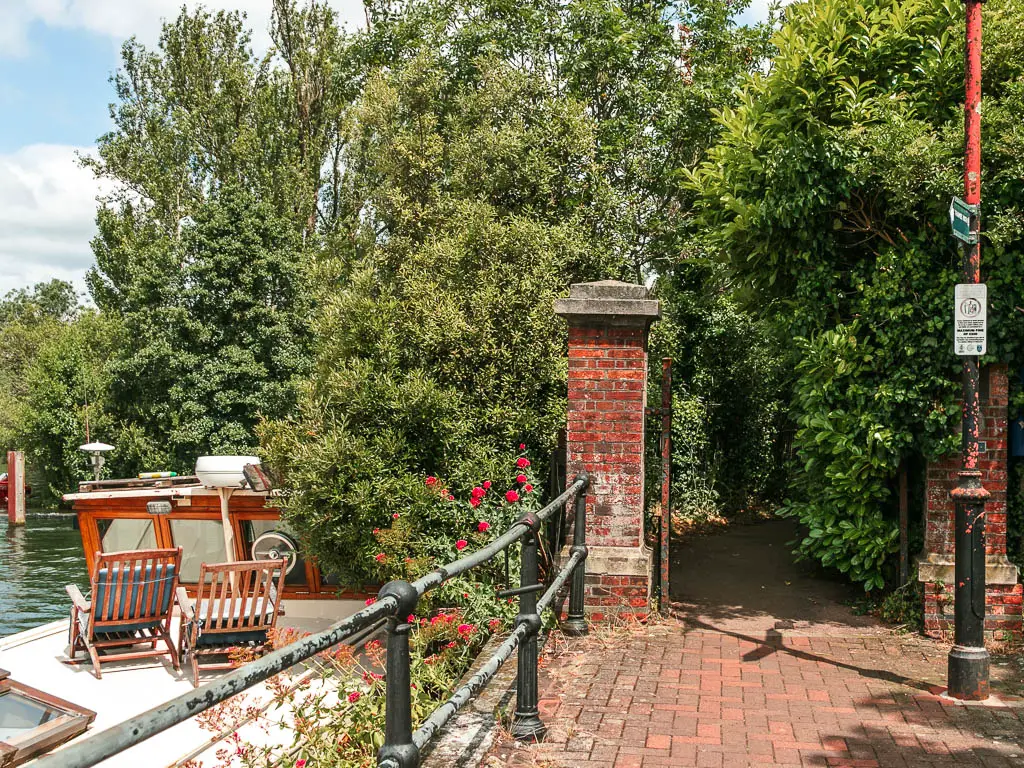 A paved path leading through two brick pillars onto a tree lined trail. There is the top of a boat visible on the river on the left with two deck chairs on top.