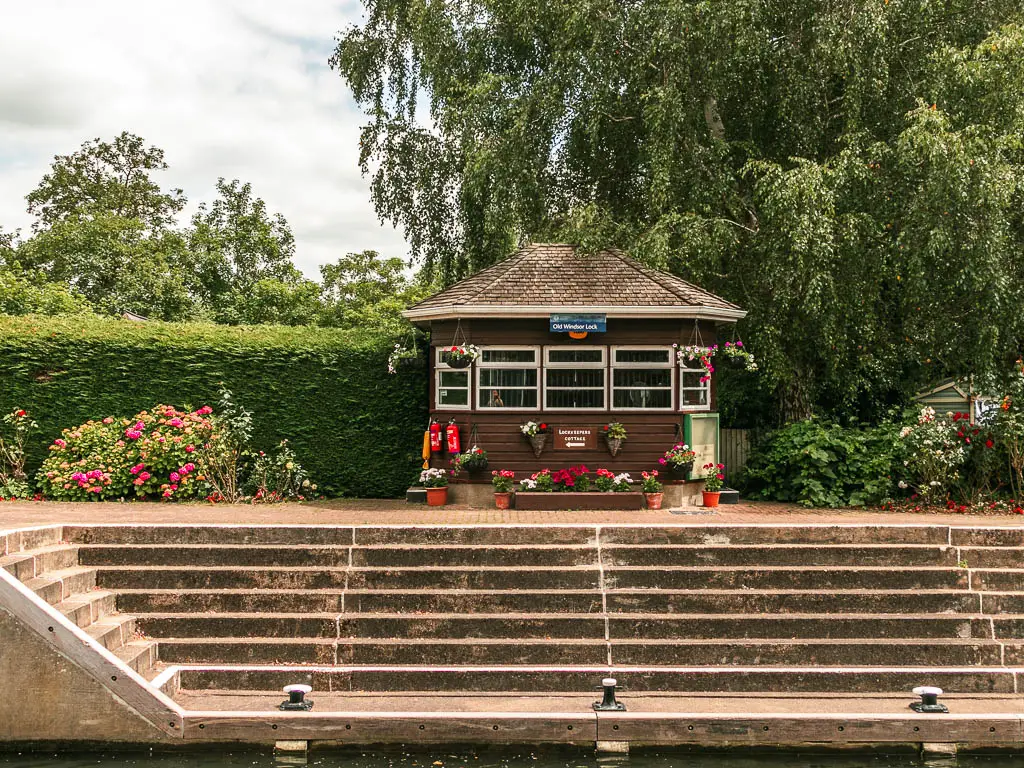 a small lock hut with flowers outside and wide steps below. There is a neatly cut hedge to the left of the hut and big tree to the right.