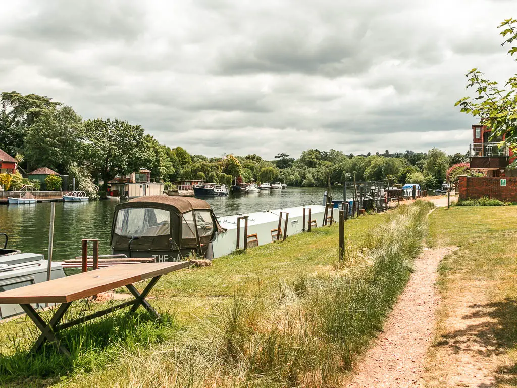 A trail leading through the grass, with the river to the left, and boats and barges moored to both sides. There is a wooden table on the grass on the left.