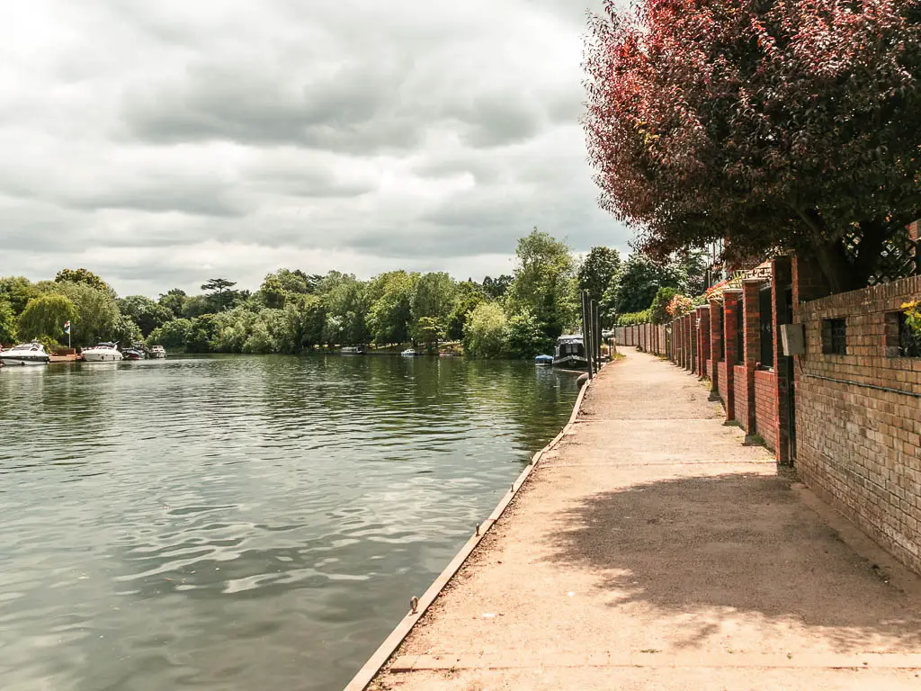 A walkway promenade leading ahead on the right, with the river to the left. There is a brick wall lining the right side of the promenaded and trees lining the river ahead in the distance. 