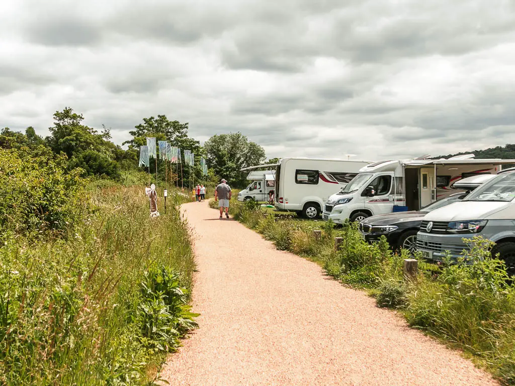 A wide path lined with tall grass and bushes to the left and parked cars to the right.