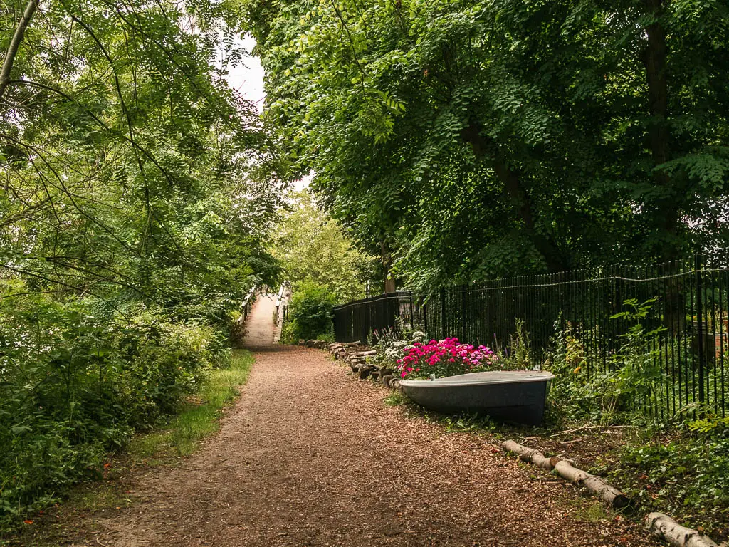 A wide trail leading towards a small bridge ahead, near the end of the walk from Windsor to Staines. There are trees and bushes on both sides of the trail. And a boat flower pot to the right.