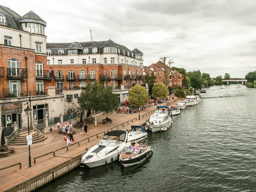 Looking down to the river with a walkway on the left side and some big buildings. There are numerous white boats moored to the side, and lots of people walking on the walkway.