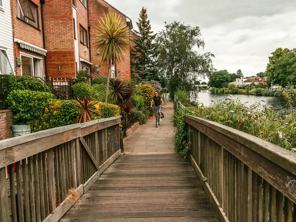 Looking along a wooden bridge to a paved path ahead, and houses on the left side and river to the right. There is a person cycling on the path. There and hedges and bushes in front of the houses.