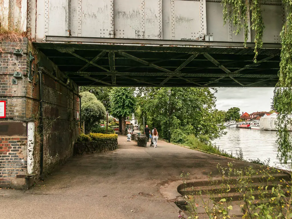 A wide path leading under a metal bridge with the river to the right, and people walking ahead.