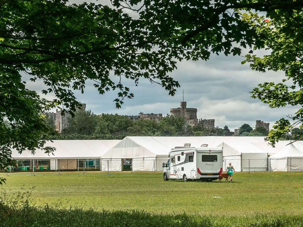 A neatly cut grass field with a big white camper van in the middle and white big tents behind, and a view to Windsor castle in the distance. There are leafy tree branches hanging into the frame from above.