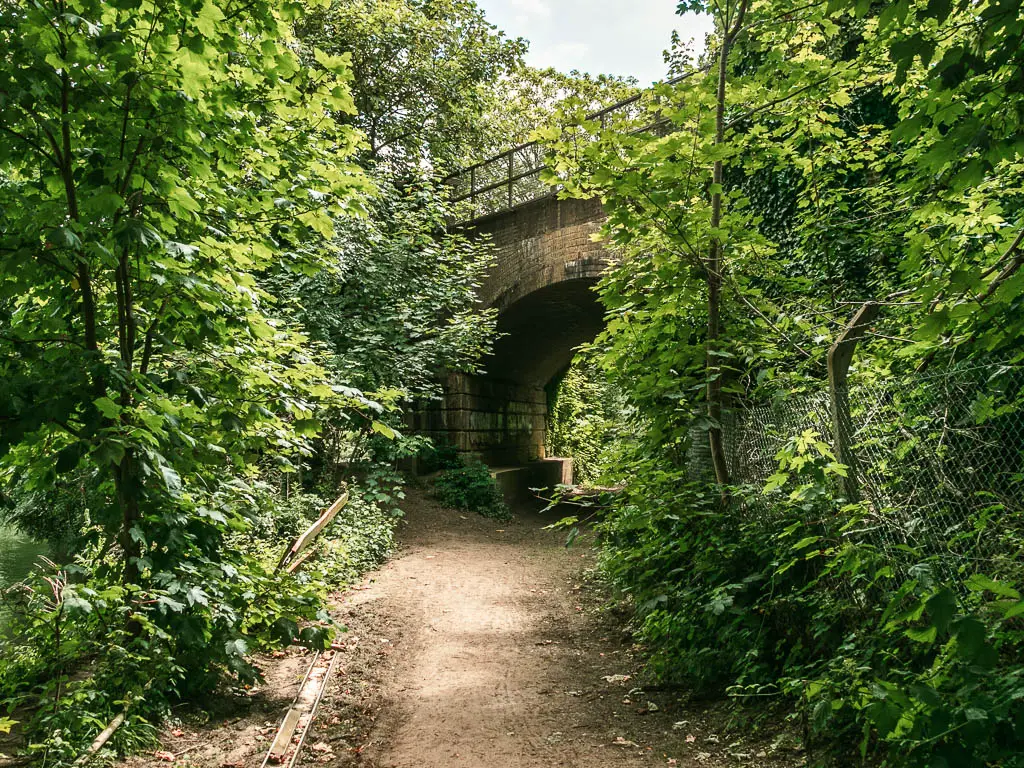 A wide dirt path lined with bushes and trees, leading under an archway bridge ahead.