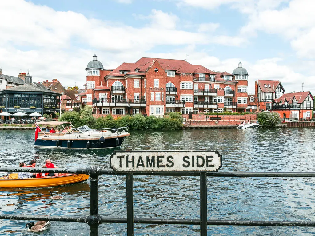 A sign saying 'Thames side' on the black railings with the river behind, at the start of the walk from Windsor to Staines. There is a small yellow boat and a bigger white boat in the choppy river. There is a big red bricked building on the other side of the river.