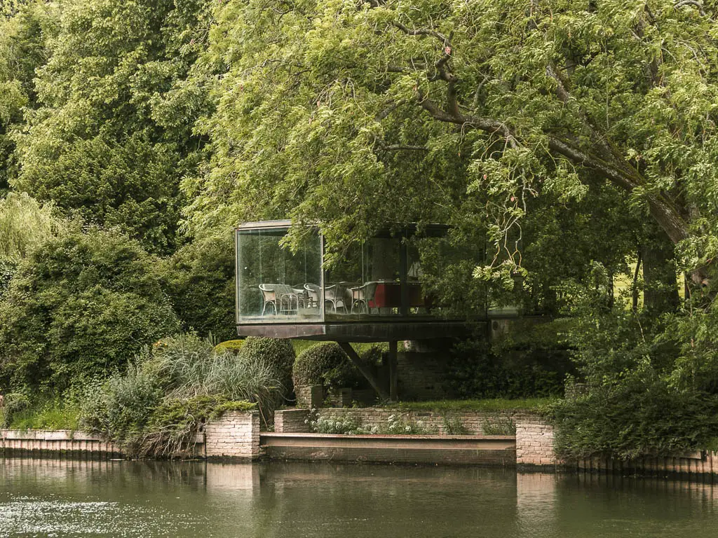 Looking across the river to a glass walled building jutting out from the trees over the water, at the start of the walk from Goring and Streatley to Reading.