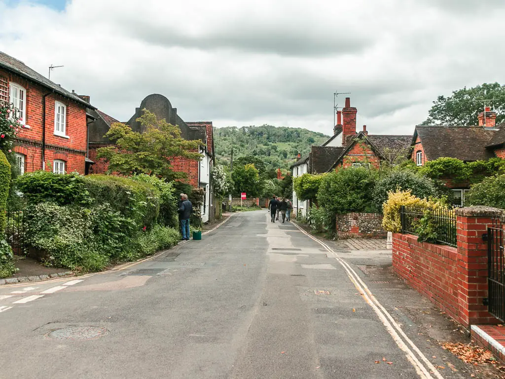 A road leading ahead, lined with residential houses, hedges, bushes, and red bricked walls. There is a big green tree covered hill ahead din the distance.