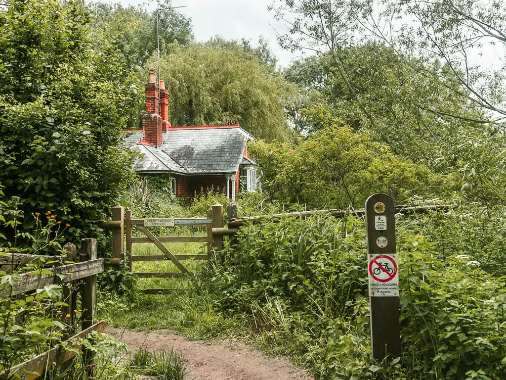 A wooden gate surrounded by bushes, with a partially hidden cottage on the other side. There is a trail curving to the left on this side of the gate, with a wooden signpost on the right side of it.