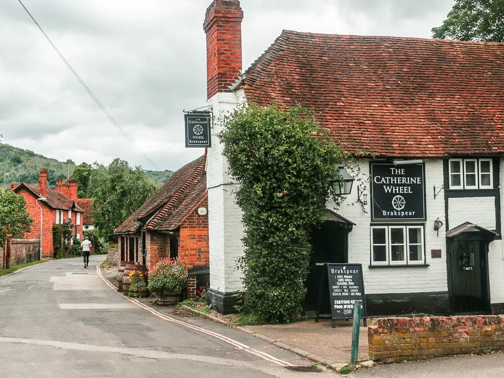 A white painted bricked walked pub building on the right, with a red brick chimney, and a bush growing up the side of it. The road is on the left.