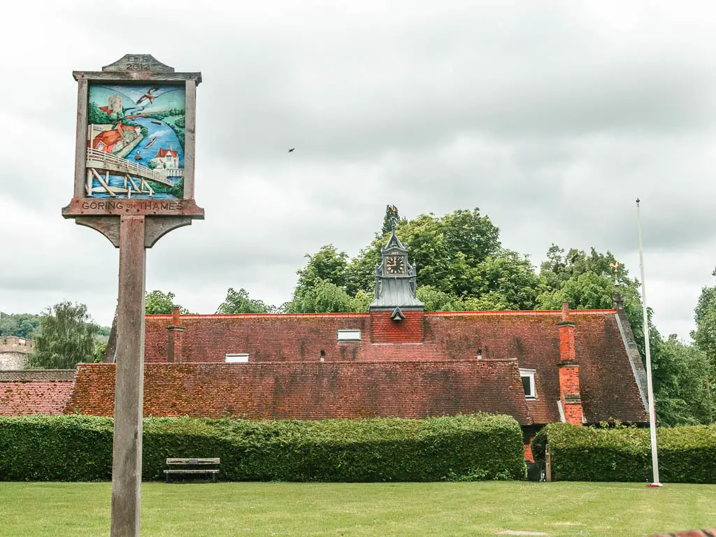 A wooden sign saying Goring and Streatley, sitting on a neatly cute green lawn, with a red roofed house on the other side behind a hedge.