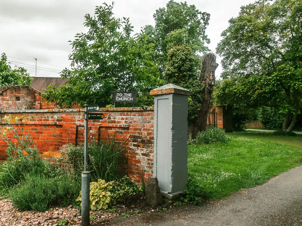 A red bricked wall on the left with grey painted pillar on the right end of it. There are plants on this side of the wall and green grass on the other side. There is a trail signpost pointing through the wall.
