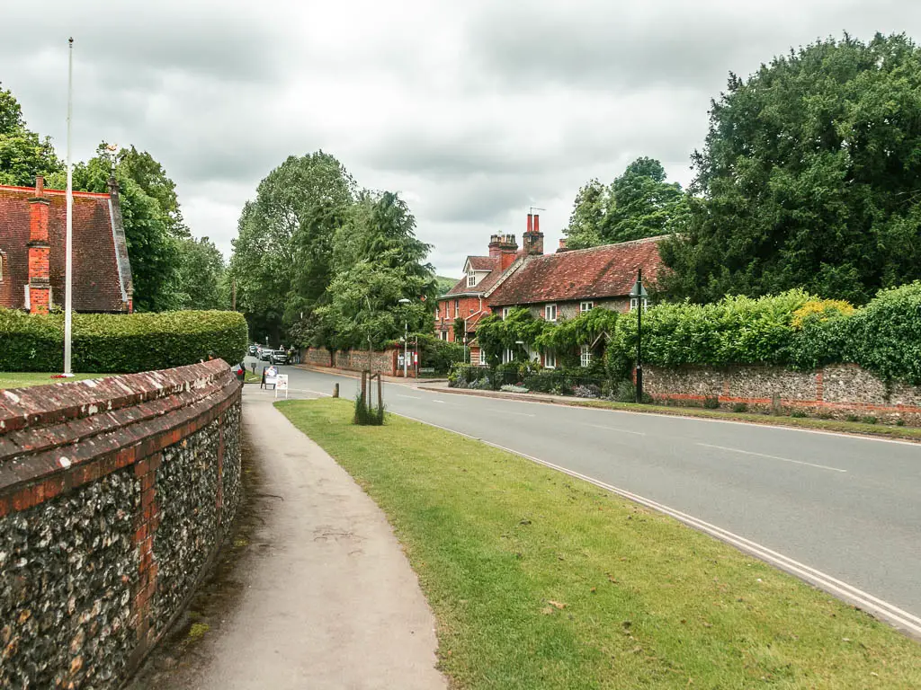 A pavement on the left with a stone wall to the left of it, and a strip of grass to the right, then the road on the right of it. There is a stone wall topped with bushes on the other side of the road, and houses covered in greenery.