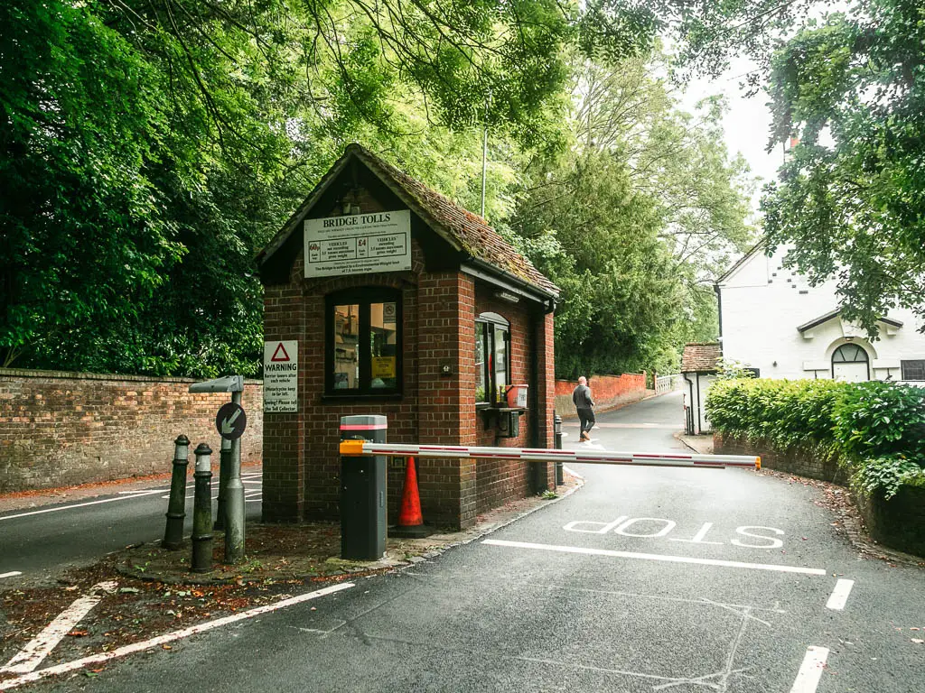 A small brick walled toll booth in between tow roads. The barrow on the right road is closed. The left road is lined with a brick wall and big trees. On the right side of the right road is a white walled house. There is a person walking on the road ahead.