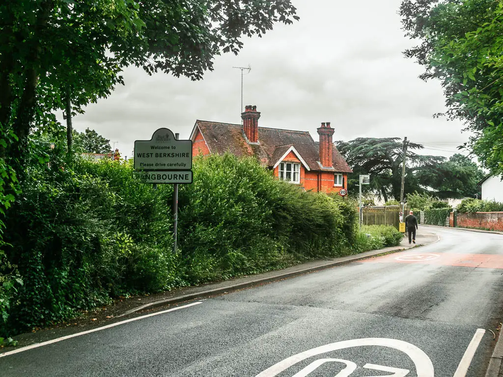 Looking across the road to a wall of green bushes, and partially hidden house behind them. There is a white sign poking out from the bushes saying 'welcome to west Berkshire'.