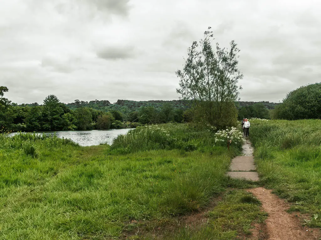 A path on the right, leading through the grass field, with the river partially visible to the left. There is a women in a white jacket walking along the path ahead.