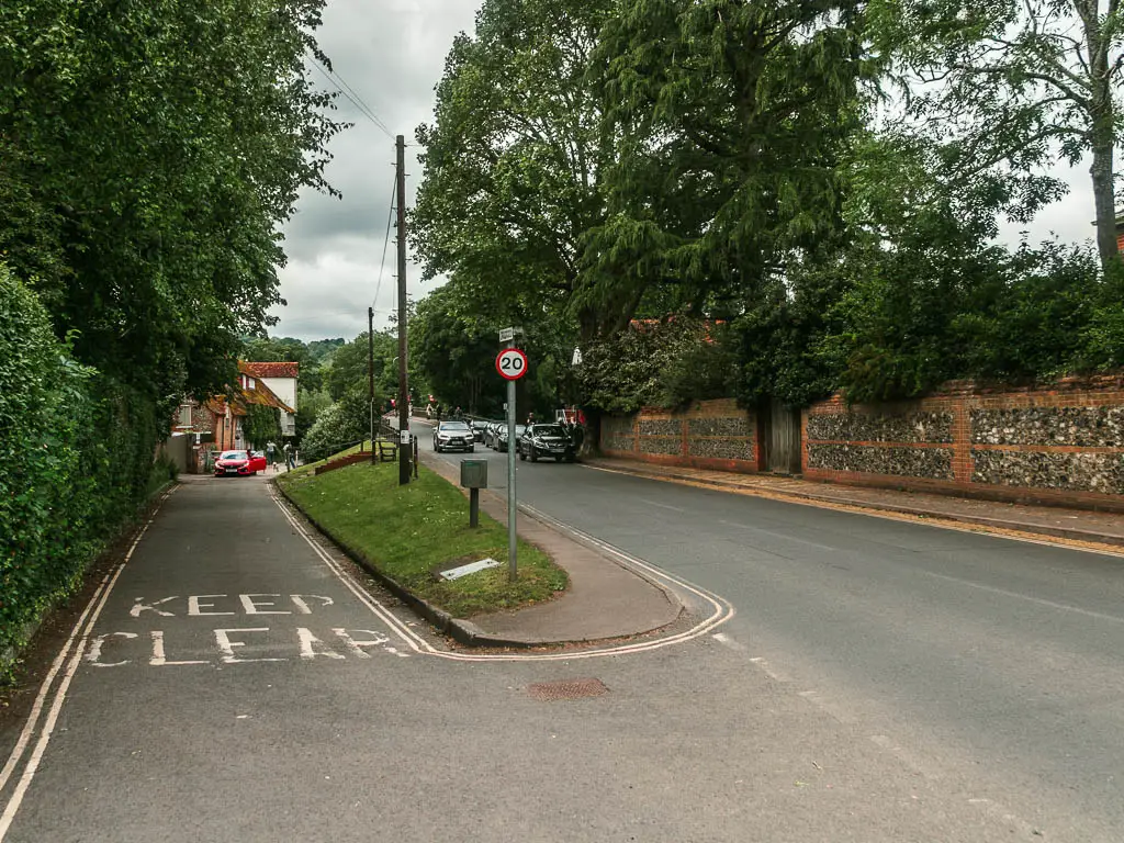 Two roads with an island separating them. The left road is lined with hedges and there is a stone wall lining the other side of the road on the right, with trees and bushes poking out from behind it.