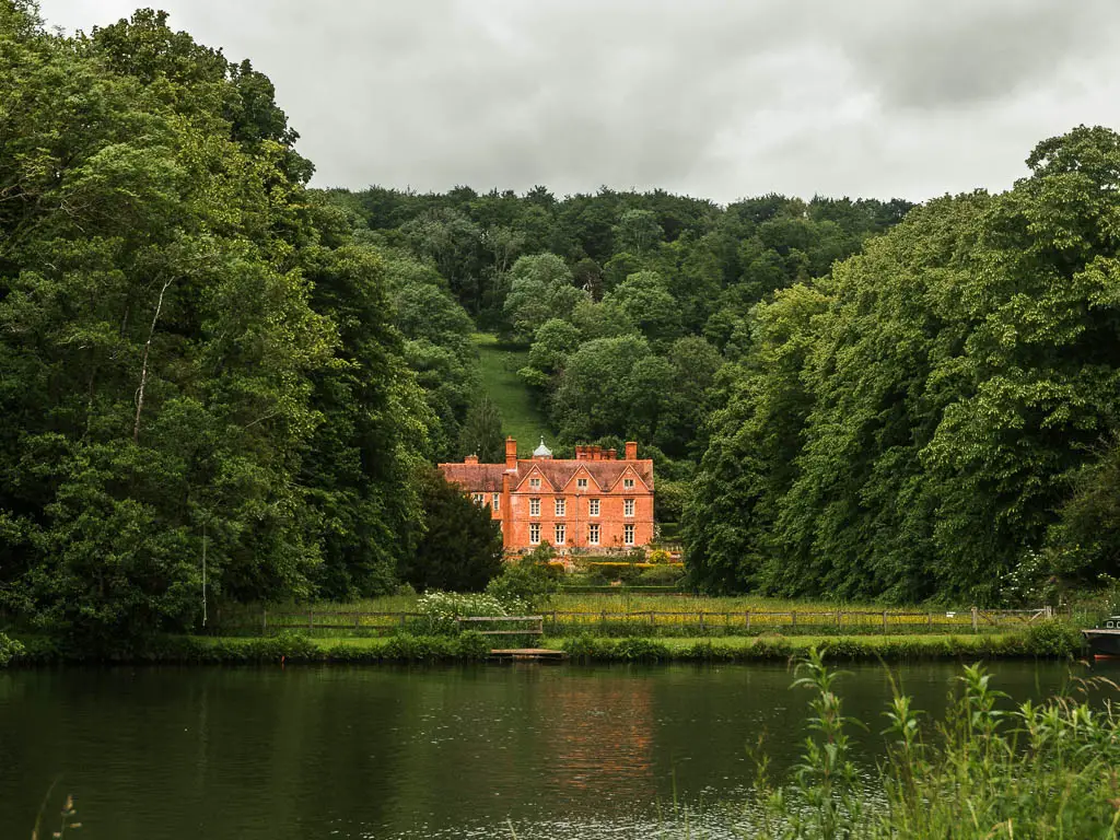 Looking across the river to a grand orange walked manor house, partway through the walk from Goring and Streatley to Reading. There is a green lawn in front of the house, and the show is surrounded by masses of tall green leafed trees.