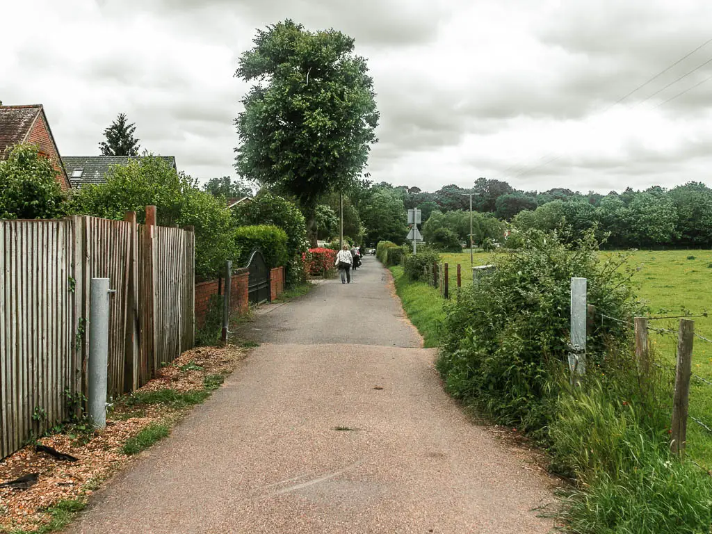 A road leading straight ahead, with a wall on the left, with rooftops poking up the top, and bushes and wire fence to the right, with a grass field on the other side. There are people walking on the road ahead.