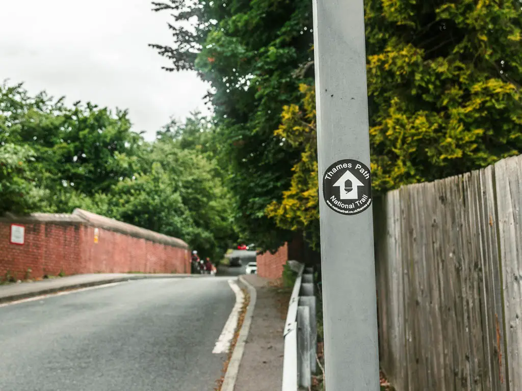A black Thames Path sticker on a metal pole, with white arrow pointing ahead along the road