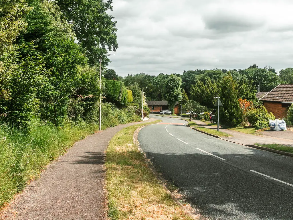 A road leading downhill, with a pavement on the left side. The left side of the pavement is lined with bushes and trees. There are houses further down the road, and trees at the bottom.