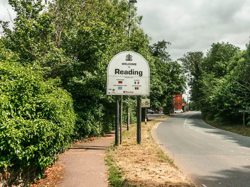 The road on the right and pavement on the left, with a big white sign saying 'welcome to reading'. The left side if the pavement is lined with green leafed bushes and trees.