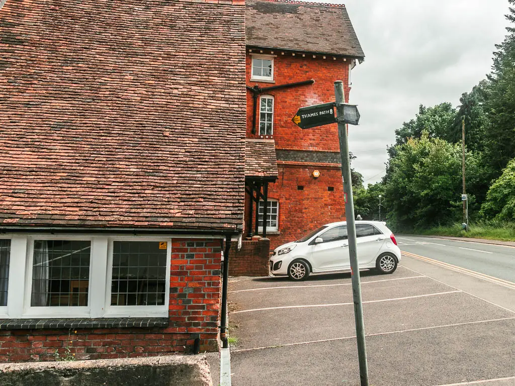 Red bricked buildings on the left with a car parking area and road to the right. There is a metal pole with trail sign pointing left. There is a white car parked next to the buildings.
