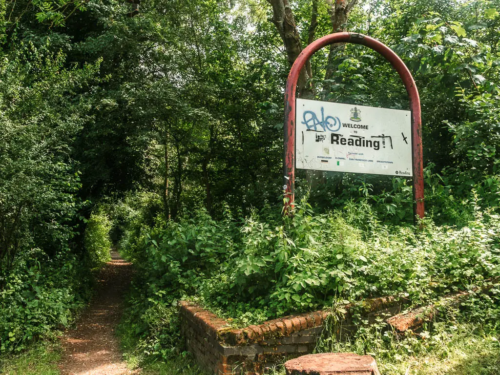 A trail on the left leading through a mass of green leafy bushes, and a big sign on the right saying 'welcome to reading;