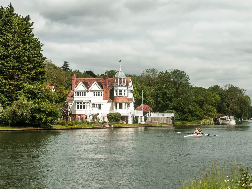 A big white walled house on the other side of the river, surrounded by trees, near the end of the walk from Goring and Streatley to Reading. There are rowers on the river.