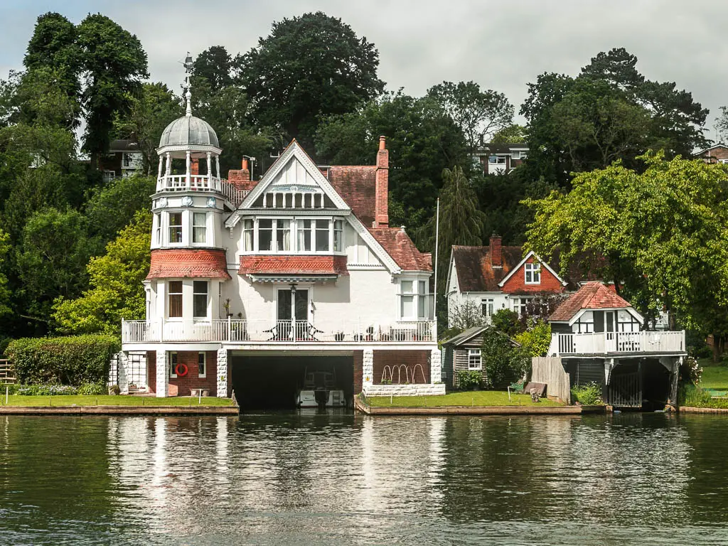Looking across the river to a large white walled house with boat garage underneath, at the end of the walk between Goring and Streatley and Reading. There are tall green leafy trees behind the house.