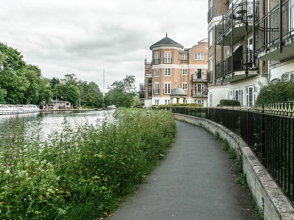 A walking path on the right, craving ahead to the left. The right side is lined with metal railings and apartment buildings. The left side is lined with tall grass and plants, with the river to the left of it.