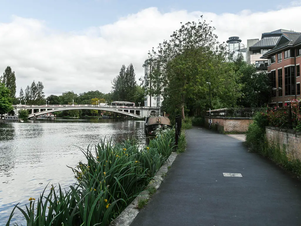 A wodę path leading ahead, with the river to the left and a bridge over it ahead. There are buildings to the right.