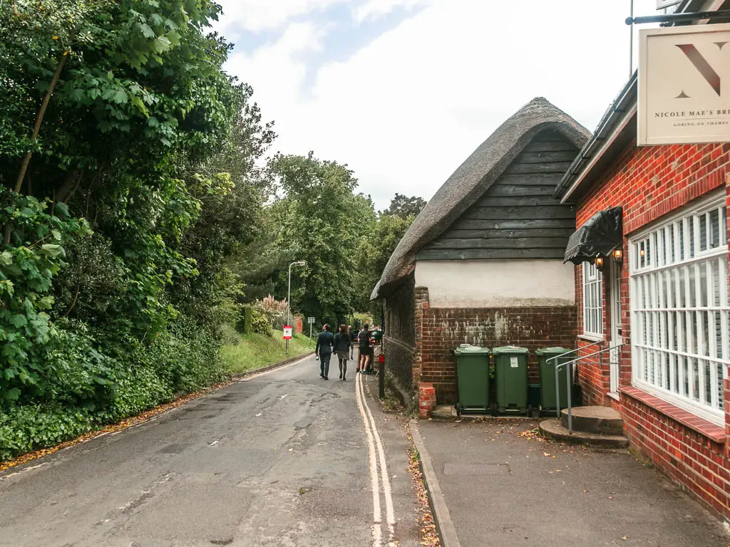A road leading ahead, with a red brick walled bustling to the right and a brown brick walled thatched roofed building next to it. There are a few people walking on the road ahead. There are green leafy bushes and trees lining the left side of the road.