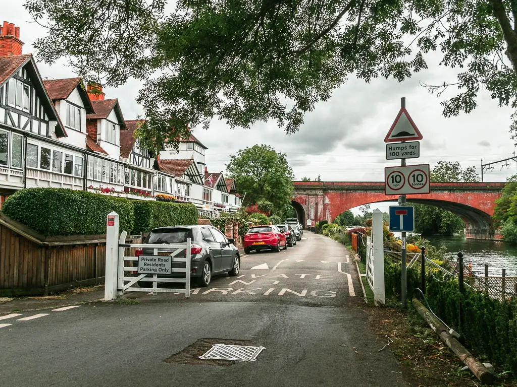 Looking along a road lined with Tudor house on the left and parked cars in front. The road leads towards a red bridge ahead.