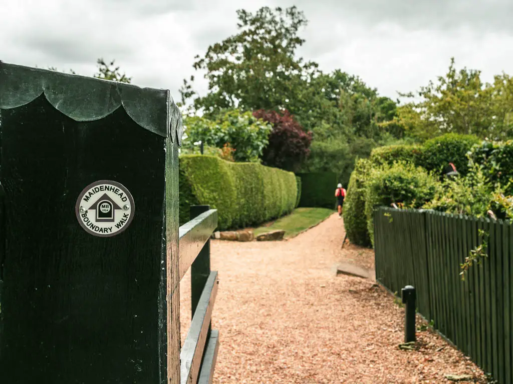 A white trail sticker on the fence wall on the left, pointing ahead along the gravel path. There are neatly cut hedges lining the path ahead.