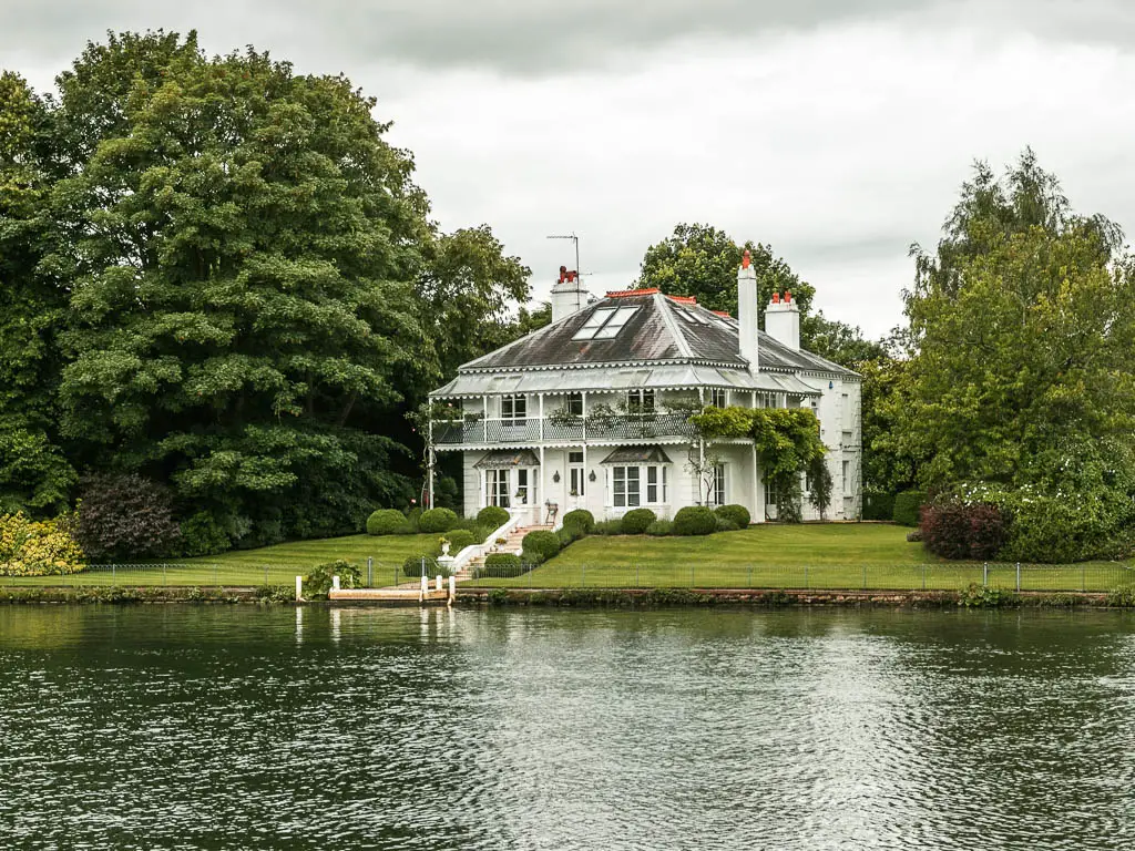 Looking across the river to a big white walked house with neatly cute green front lawn, on the walk from Maidenhead to Windsor. The hose is surrounded by big leafy trees.