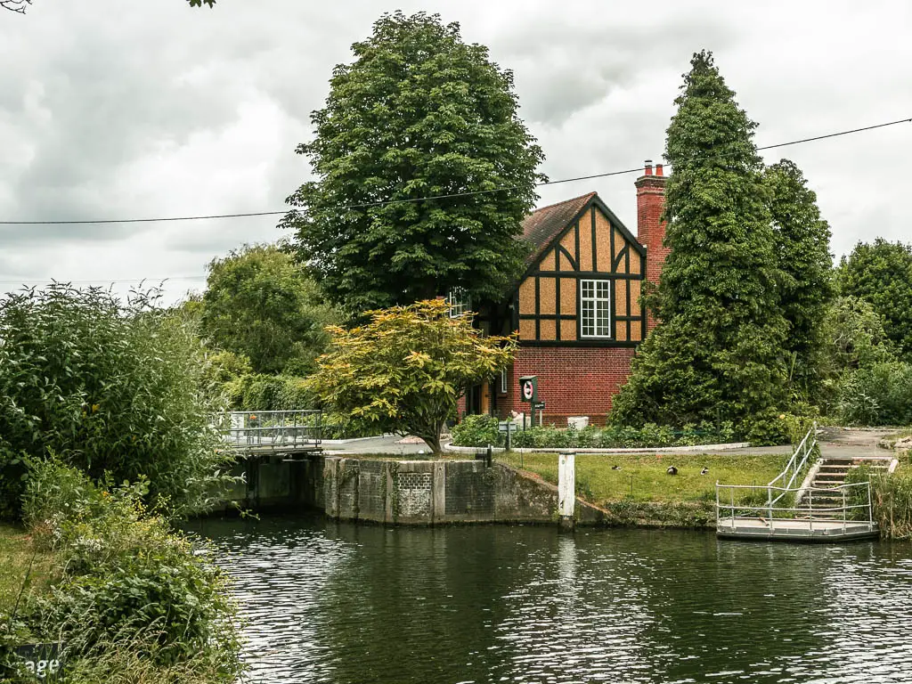 Looking across the river to a partially hidden Tudor style lock house, surrounded by different types of trees, on the walk between Maidenhead and Windsor.