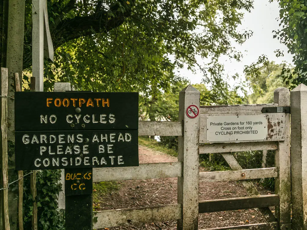 A sign on the left saying 'footpath, no cycles', with a dirty white wooden gate and fence behind it.